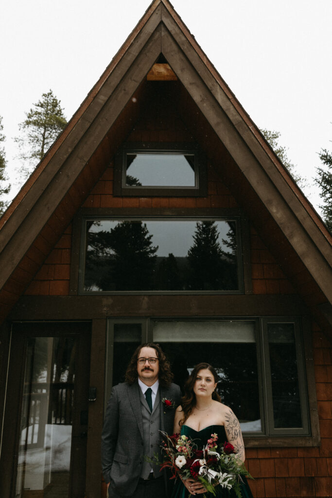 A bride and groom stand in front of A Frame cabin in alternative wedding gear.  