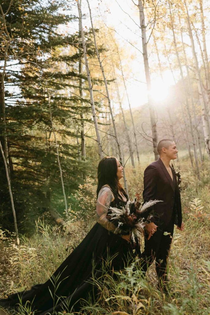 A lgbtq couple smiles while looking at their officiant prior to their wedding ceremony in the Colorado mountains.  A bride wears a black dress and holds a textured bouquet while her partner wears a plum colored suit.