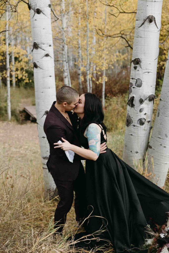 A lgbtq couple smiles while looking at their officiant prior to their wedding ceremony in the Colorado mountains.  A bride wears a black dress and holds her partner who wears a plum colored suit.