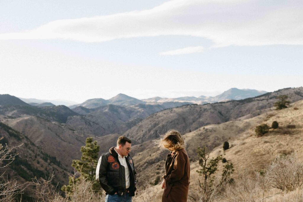 A newly engaged couple stands on a mountain with a beautiful landscape of mountains and valleys behind them in Colorado.  