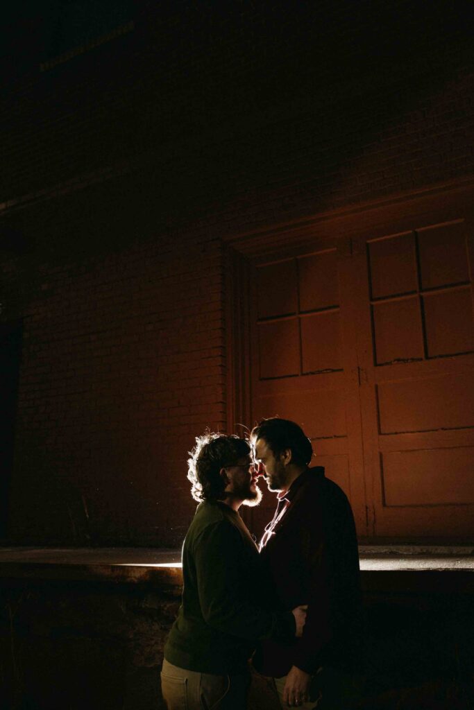 Two men stand almost kissing with a light shining behind them in downtown Denver during their fall engagement session. 